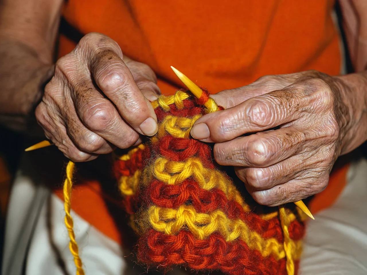 Primo piano di mani anziane che lavorano a maglia con lana color terracotta e senape.