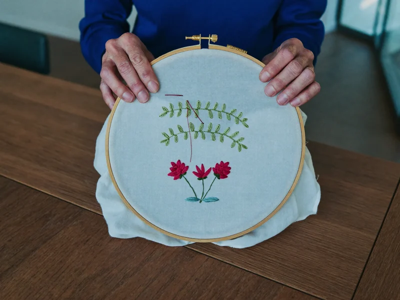 Mani anziane che ricamano fiori su lino teso in un telaio di legno.